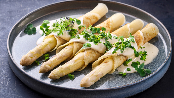 KI generiert: Weißer Spargel in Pfannkuchen gerollt, mit Sauce und frischen Kräutern auf einem Teller angerichtet.