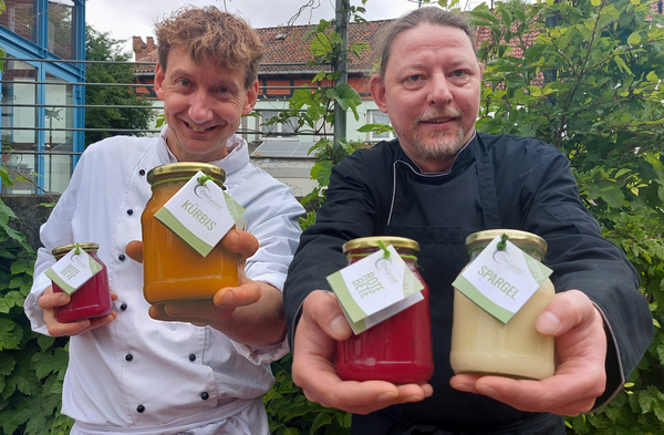 KI generiert: Zwei Köche halten Gläser mit Suppen, beschriftet mit "Kürbis", "Rote Bete", "Spargel".