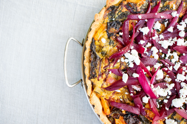 Eine Quiche mit Karotten in einer Backform, umgeben von Eiern, Paprika, Mehl und Kräutern.
