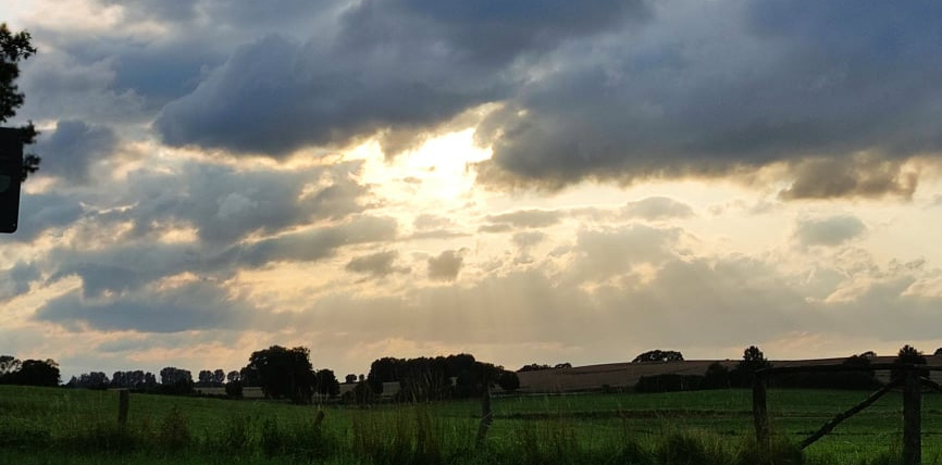 Eine Landschaft mit grünen Feldern und dramatischen Wolken am Himmel bei Sonnenuntergang.