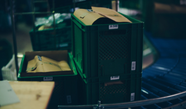 KI generiert: Grüne Plastikkisten auf einem Förderband, mit Papiertüten bedeckt. Labels: "A-311", "A-348".