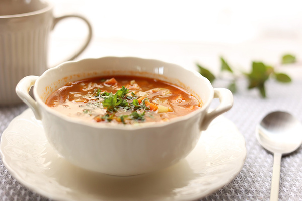 KI generiert: Das Bild zeigt eine weiße Suppenschüssel mit einer würzig aussehenden Gemüsesuppe, garniert mit frischen Kräutern. Im Hintergrund sind eine Tasse und ein Löffel zu sehen.