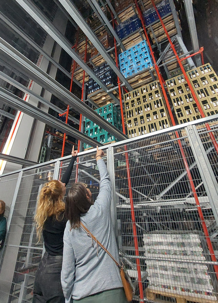 KI generiert: Zwei Personen zeigen auf Getränkekisten in einem hohen, regalen Lagerhaus.