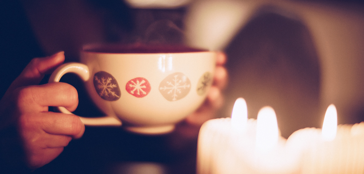 KI generiert: Eine Hand hält eine dampfende Tasse mit Schneeflockenmotiv, im Hintergrund sind Kerzen zu sehen.