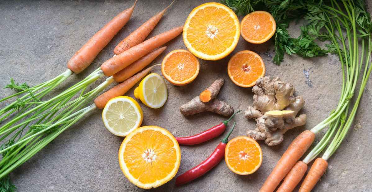 Das Bild zeigt eine Zusammenstellung frischer Zutaten: Karotten, Orangenscheiben, Zitronen, Chilischoten, Ingwer und Kurkuma auf einer Steinoberfläche. Der Fokus liegt auf der Vielfalt an Gemüse und Früchten.