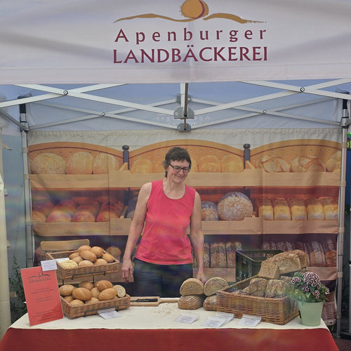KI generiert: Eine Frau steht hinter einem Bäckereistand mit verschiedenen Broten. Text: "Apenburger LANDBÄCKEREI".