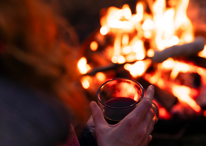 Das Bild zeigt eine Person, die eine Tasse wärmt, während im Hintergrund ein Lagerfeuer lodert. Der Fokus liegt auf der gemütlichen Atmosphäre und der Wärme des Feuers.