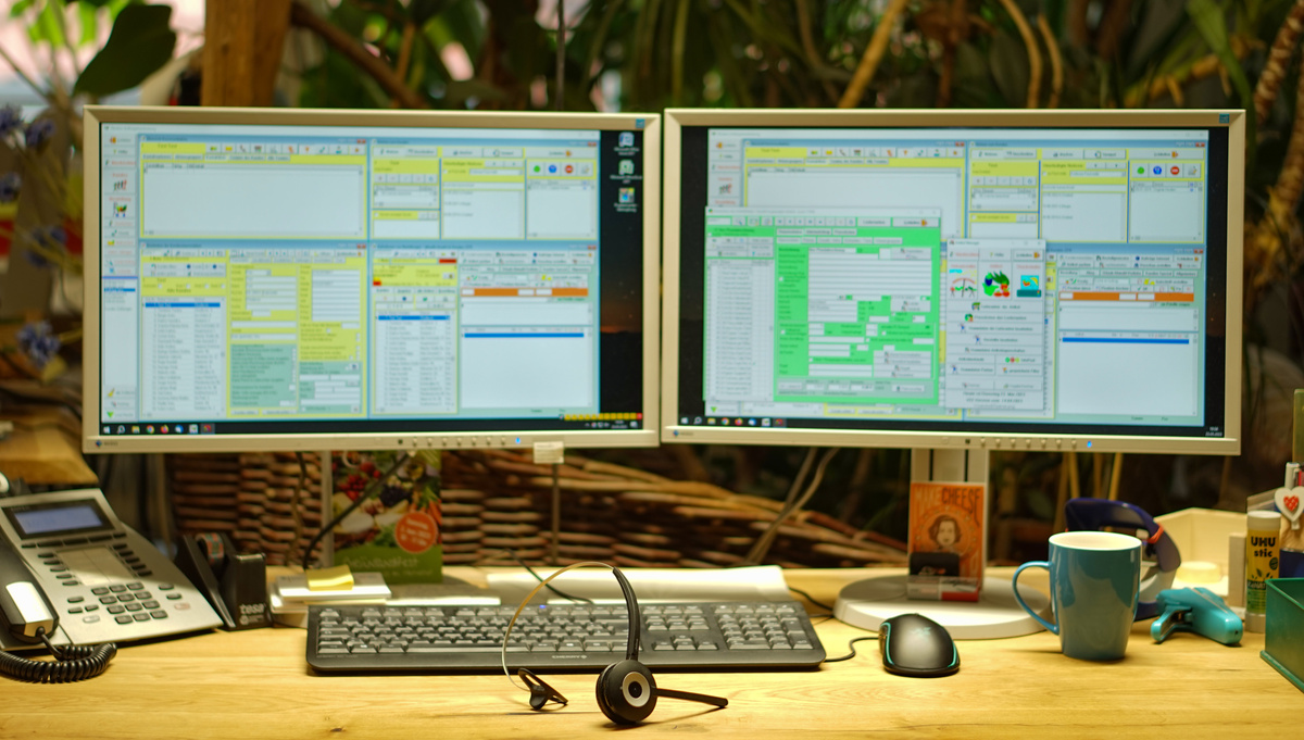  Zwei Bildschirme mit komplexen Software-Oberflächen, Telefon, Tastatur und Headset auf einem Schreibtisch