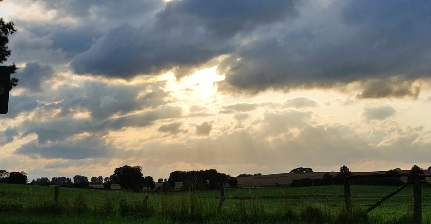 KI generiert: Wolkenverhangener Himmel mit Sonnenstrahlen über einer grünen Wiese und vereinzelten Bäumen.