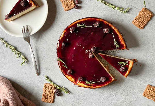KI generiert: Das Bild zeigt einen Käsekuchen mit einer glänzenden roten Glasur, garniert mit Beeren und Rosmarinzweigen. Ein Stück wurde herausgeschnitten und liegt auf einem separaten Teller; Spekulatiuskekse sind dekorativ um den Kuchen arrangiert.