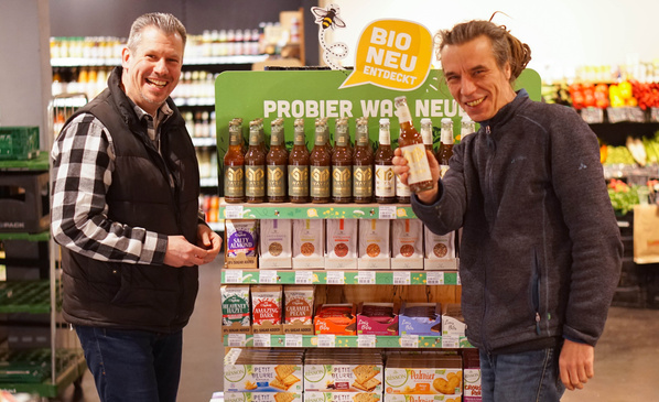 KI generiert: Zwei Männer stehen vor einem Regal mit Bio-Produkten. Text: "PROBIER WAS NEU" und "BIO NEU ENTDECKT".