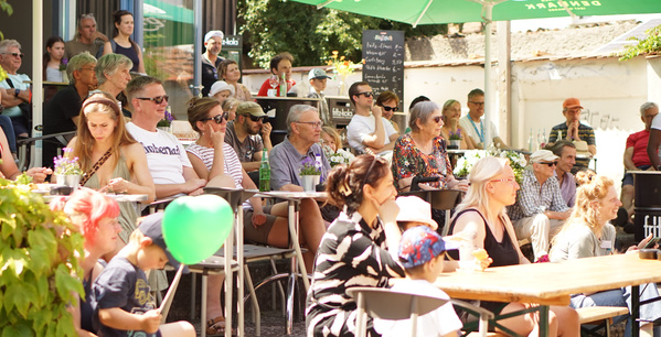 KI generiert: Eine Gruppe von Menschen sitzt draußen bei einem Event. Im Hintergrund steht eine Tafel mit Menütext.