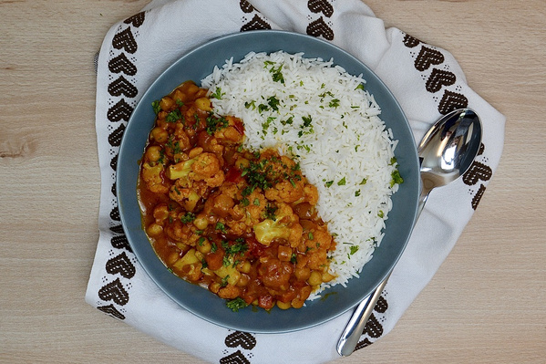 KI generiert: Ein Teller mit Curry und Reis, dekoriert mit Kräutern, daneben ein Löffel auf einer bestickten Serviette.