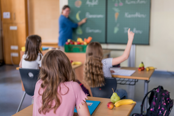 KI generiert: Schüler im Unterricht über Früchte. Auf der Tafel: Apfel, Banane, Birne, Karotte, Trauben.