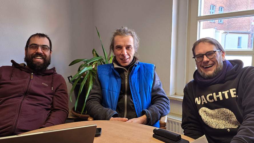 KI generiert: Drei Männer sitzen lachend an einem Tisch in einem Raum mit Pflanzen und Fenster.