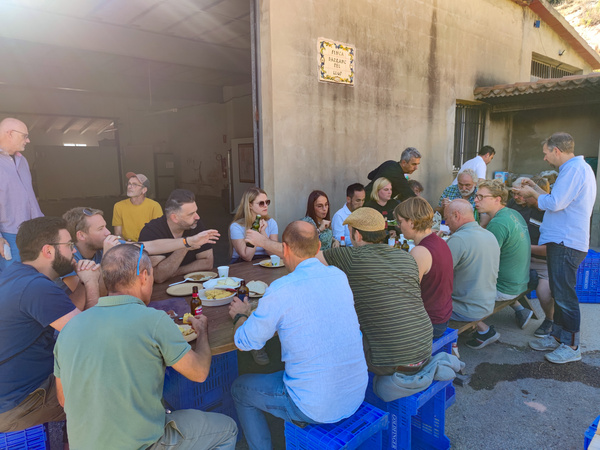 KI generiert: Eine Gruppe von Menschen sitzt an einem großen Tisch im Freien und genießt gemeinsam eine Mahlzeit. Sie scheinen sich unter freiem Himmel in einer entspannten Atmosphäre zu unterhalten.