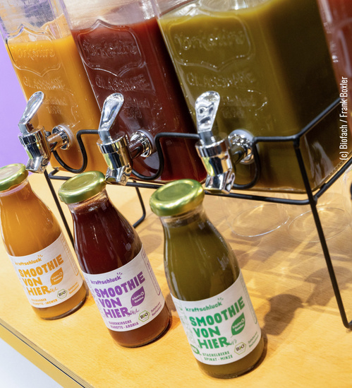 KI generiert: Drei Smoothie-Flaschen vor Getränkespendern. Aufschrift: "SMOOTHIE VON HIER".