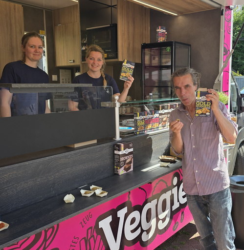 KI generiert: Drei Personen an einem Essensstand präsentieren Verpackungen von veganen Produkten. Text: „Veggie“.