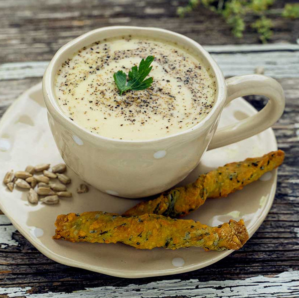 KI generiert: Eine Tasse Suppe mit Petersilienblatt, Brotsticks und Sonnenblumenkernen auf einem Teller.