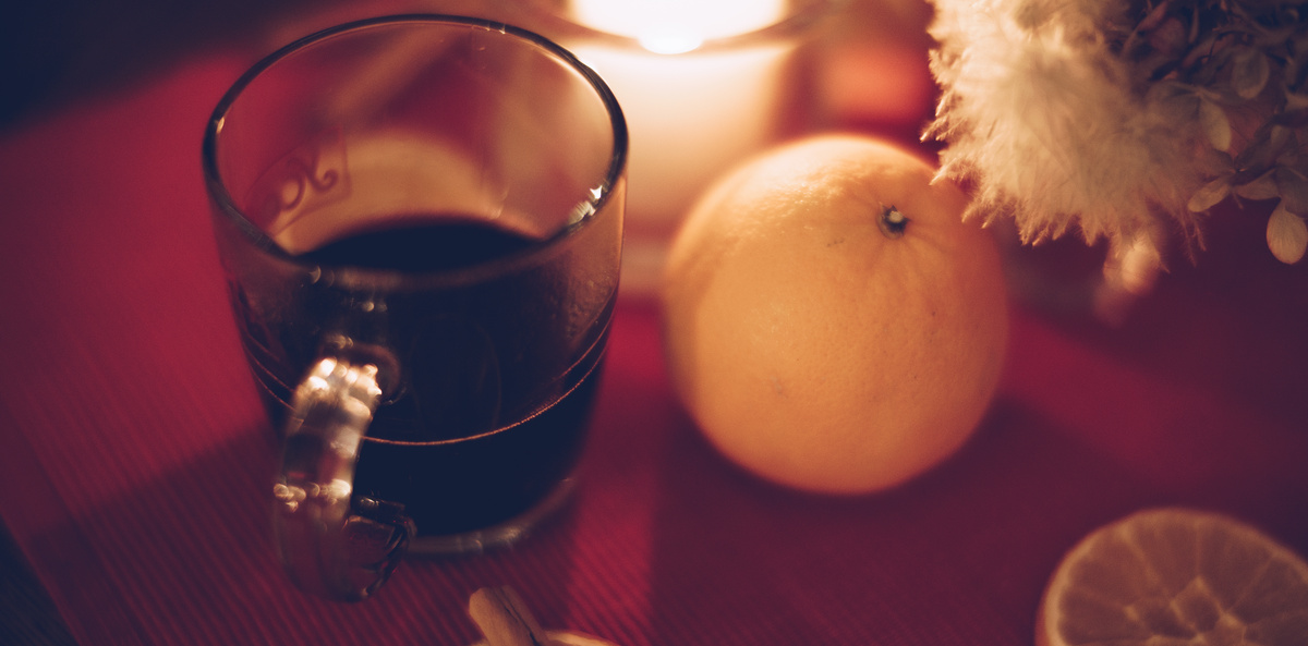 KI generiert: Ein Glas mit heißem Getränk, Orange, Kerze und Blumen auf rotem Hintergrund.