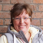 KI generiert: Eine Person lächelt vor einer Ziegelsteinwand in die Kamera und trägt einen Schal. Die Stimmung wirkt freundlich und warm.