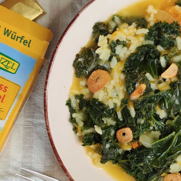 KI generiert: Ein Teller Risotto mit Grünkohl und Knoblauch. Verpackung für Gemüsebrühe ist sichtbar. Text: "Würfel" und "Gemüse".