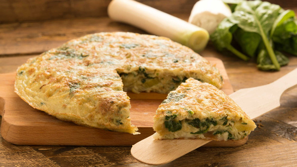 Das Bild zeigt eine frische Tortilla mit Spinat auf einem Holzbrett, von der ein Stück herausgeschnitten wurde. Im Hintergrund sind Zutaten wie frisches Gemüse zu sehen.
