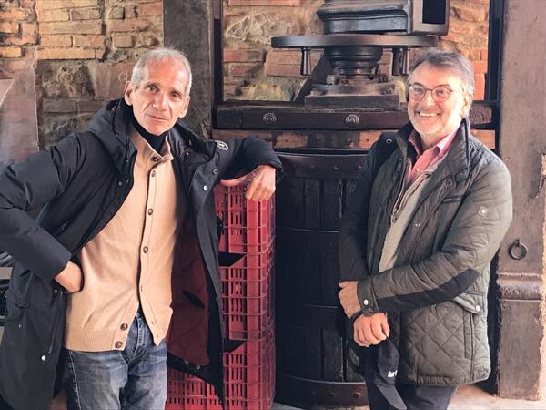 KI generiert: Zwei Männer posieren neben einer Weinpresse in einem rustikalen, Backsteingebäude.