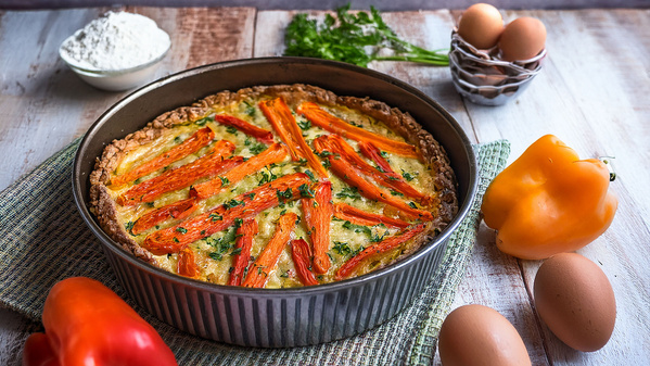 Eine Quiche mit Karotten in einer Backform, umgeben von Eiern, Paprika, Mehl und Kräutern.