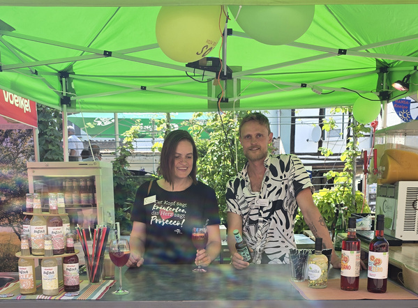 KI generiert: Zwei Personen an einem Getränkestand mit bunten Flaschen und Straws. Text auf T-Shirt: „Der Kopf sagt Elternzeit...“