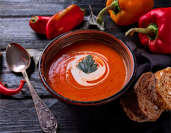 KI generiert: Eine Schüssel mit roter Suppe, garniert mit Sahne und Petersilie, umgeben von Paprika und Brot.